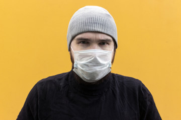 a man in a black jacket wearing a disposable mask to protect against coronavirus. Yellow background, emotions on the face. Person in a grey hat. Safety measures in case of illness.