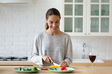 Smiling young woman cutting vegetables for salad on wooden board, using knife, standing in modern kitchen with red wine glass, happy female preparing food for dinner or party alone - Powered by Adobe