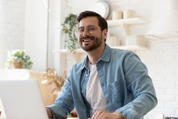 Happy young man wearing glasses laughing, using laptop, excited by good news, successful smiling...