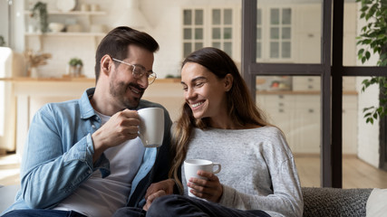 Happy loving couple relaxing on couch, drinking hot beverages, tea or coffee, holding white cups,...
