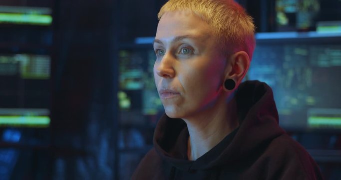 Close up of female Caucasian hacker working on computer in front of big screen and turning face to camera in dark room. Woman committing cyber crime and taking on hood. Internet data security concept.