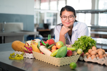 Portrait black long hair Asian pretty young woman nutritionist smiling with out of focus vegetables, fruits and eggs in the kitchen.The chef is thinking of creating a menu.