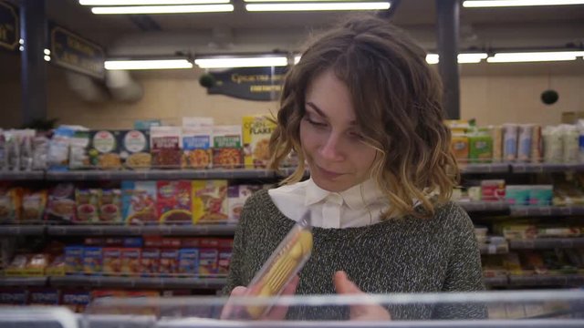 Young curly haired woman doing grocery shopping at the supermarket, she is reading a product label and nutrition facts on a box with cheese. Footage from the shelf