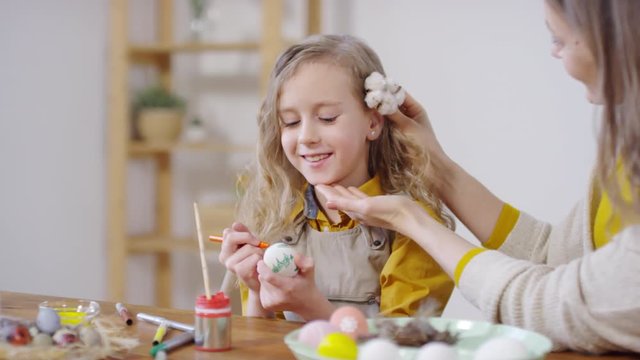 Handheld of mother smiling and putting cotton flower behind ear of cheerful little girl sitting at table and drawing on Easter egg
