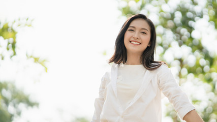 Portrait of young Asian woman wearing the white shirt in casual style smiling and enjoy life in the natural sunlight in the park, Relaxing moment, enjoying the nature, mindful and feel the fresh are.