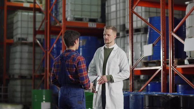 Tracking medium shot of middle aged enginner in white coat giving instructions to young warehouse worker with clipboard in factory storehouse