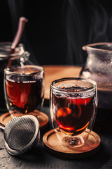Cup of freshly brewed black tea, dark moody, tea ceremony. Hot water is poured from the kettle into a flass cup on wooden tray, black concrete background.