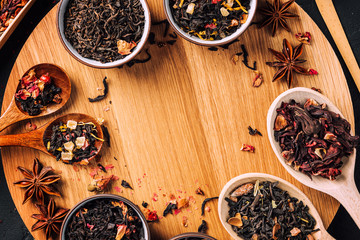 Assorted dried tea leaves in bowls and wooden spoons on a concrete black background. Dark moody. Set of different herbal and fruit tea