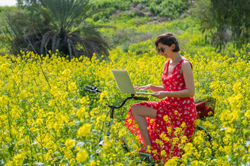 a freelancer girl works behind a laptop in a spring flower and sunny field. a freelancer girl is working at a laptop in a spring blooming and Sunny field. the girl came to the field on a Bicycle and