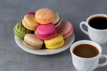 Assorted macaroon cakes in a white plate and two white cups of coffee on a gray background.