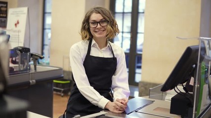 Woman in black apron as a cashier at the cash register in the supermarket or discounter. Smiling short haired curly woman smiling to the camera