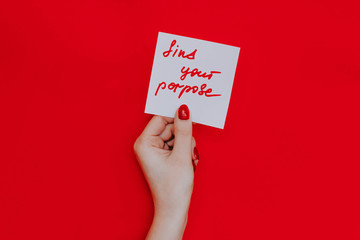 Note in a female hand with manicure, red nails. "find your purpose" sign. Background red