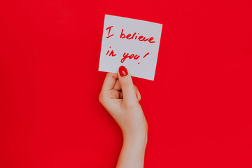 Note in a female hand with manicure, red nails. "I believe in you!" sign. Background red