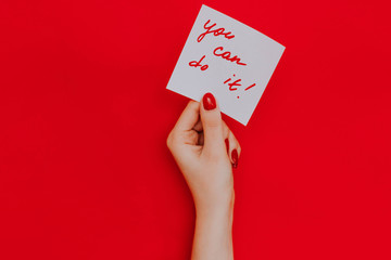 Note in a female hand with manicure, red nails. "You can do it!" sign. Background red