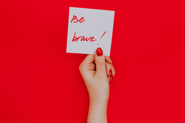 Note in a female hand with manicure, red nails. "Be brave!" sign. Background red