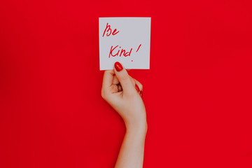 Note in a female hand with manicure, red nails. "Be kind!" sign. Background red