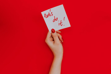 Note in a female hand with manicure, red nails. "Let's do it!" sign. Background red