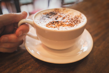 Cappuccino in a white cup and saucer on a wooden table with a hand holding the handle. Shallow focus.