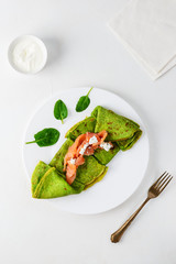 Thin spinach pancakes with pieces of salmon and curd cheese on a white plate. Healthy food concept, gluten free.