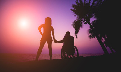 Disabled man in a wheelchair with his wife on the beach. Silhouettes at sunset