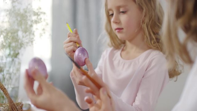 Handheld shot of happy young woman and cute girl talking and putting glitter on purple Easter eggs