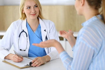 Woman doctor and patient sitting and talking at medical examination in hospital or clinic. Blonde therapist cheerfully smiling. Medicine and healthcare concept