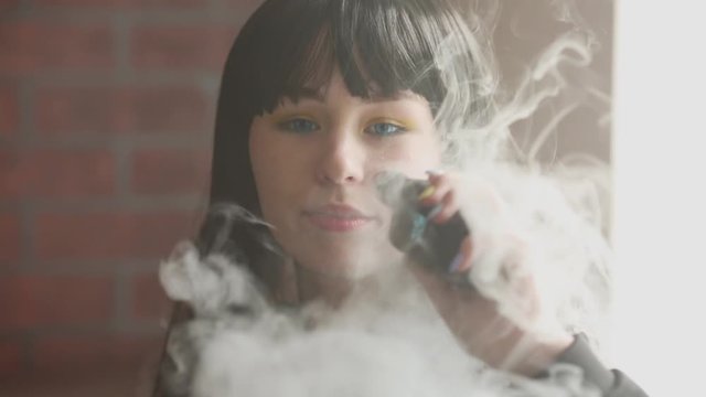 Vaping teenager. Young pretty white caucasian teenage brunette girl with blue eyes and problem skin smoking an electronic cigarette indoors. Deadly bad habit. Vape activity.