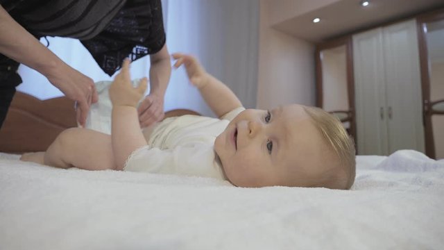 Mother and toddler concept of a happy childhood. Careful mother changes baby diaper at home. Cute toddler with blue eyes lies on a diaper in bedroom. Low-angle shot in close-up