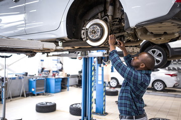 African male car mechanic repairing a car standing under the car located on the lift in the service...