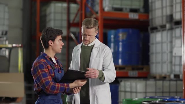 Middle aged engineer in white coat meeting young warehouse worker and checking his notes in factory storage