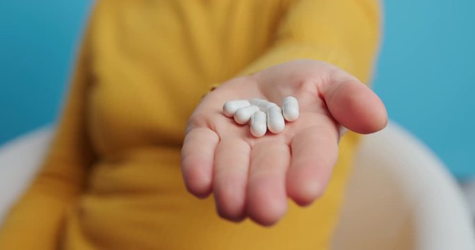 Woman's hand pours out white pills from transparent jar into palm outstretched forward to camera on blue background. Health, complex of vitamins for people. Health care. Taking medication, pills