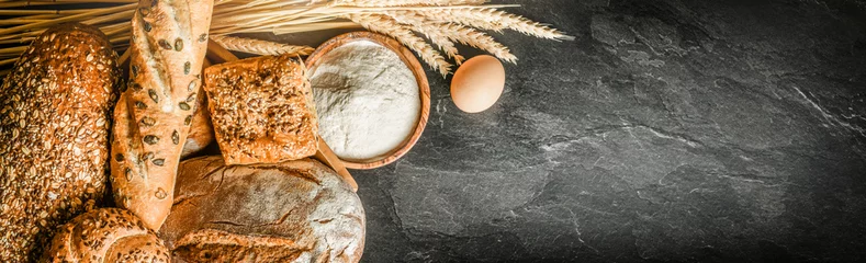 Gordijnen Brood met tarwe en een kom meel op een donker bord, het panorama van het witte bakkerijvoedselconcept of een brede bannerfoto. © Milan