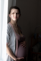 A cute pregnant young woman is looking through a curtain through a sunny window. A pregnant girl in a nightie is standing near the window and smiling.