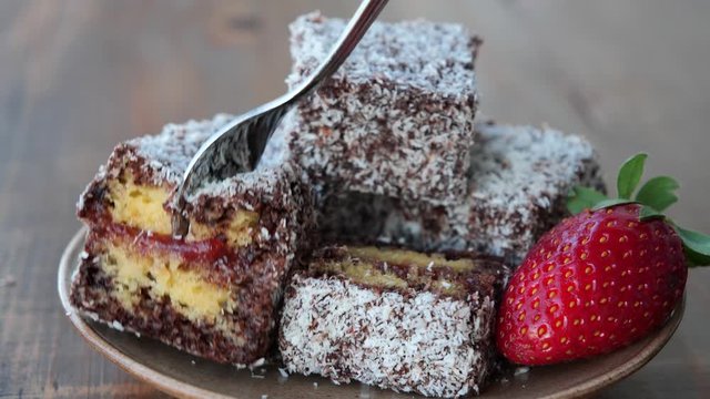 Piece of lamington cake with strawberry jam is picked up with a fork. Squares of sponge cake coated in chocolate sauce and rolled in desiccated coconut on plate