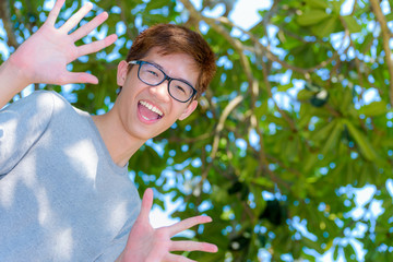 Low angle shot handsome Asian teenage boy wearing glasses fun, Portrait cheerful hipster young man are looking and laugh with a happy face at park on green nature background in Thailand