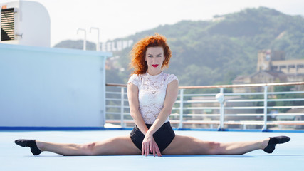 Slim beautiful girl dancer on the observation deck does gymnastic twine. red-haired curly-haired European girl on a blue podium sits in a transverse twine in sun rays on background of city. emotions