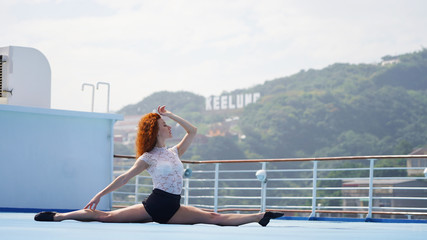 Close-up of beautiful flexible ballerina with red hair sitting on a twine against a blue background. A bright curly girl sitting in a split profile mysteriously looks up on the deck of a cruise ship.