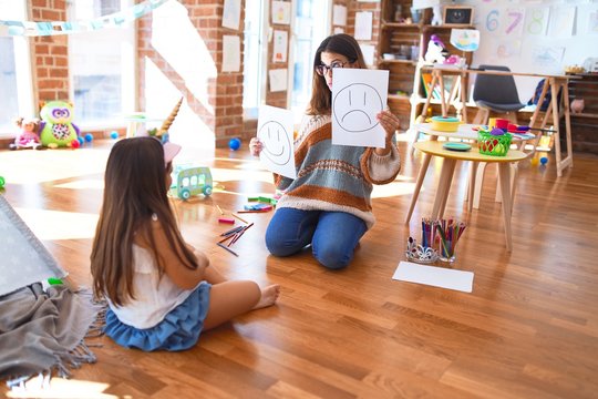 Beautiful pshychologist and toddler doing therapy with emotions emoji around lots of toys at kindergarten