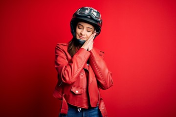 Young beautiful brunette motocyclist woman wearing motorcycle helmet and red jacket sleeping tired dreaming and posing with hands together while smiling with closed eyes.