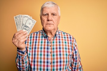 Senior handsome hoary man holding bunch of dollars banknotes over yellow background with a confident expression on smart face thinking serious