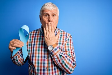 Senior handsome hoary man holding blue cancer ribbon symbol over isolated background cover mouth with hand shocked with shame for mistake, expression of fear, scared in silence, secret concept