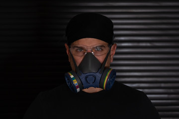 Portrait of a man with a breathing mask and safety glasses dressed in black looking at camera on dark background