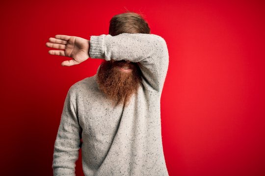 Handsome Irish redhead man with beard wearing casual sweater and glasses over red background covering eyes with arm smiling cheerful and funny. Blind concept.