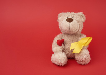 Cute brown teddy bear holds a silk yellow ribbon in the shape of a loop on a red background, concept of the fight against childhood cancer