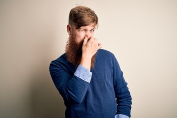 Handsome Irish redhead business man with beard standing over isolated background smelling something...