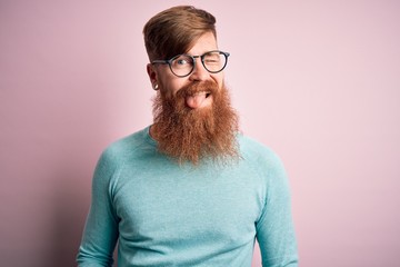 Handsome Irish redhead man with beard wearing glasses over pink isolated background winking looking...