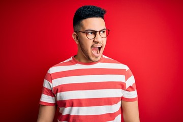 Young handsome man wearing casual striped t-shirt and glasses over isolated red background angry...