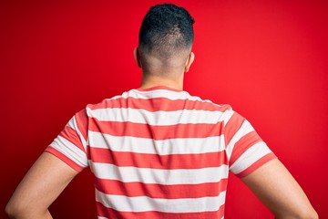 Young handsome man wearing casual striped t-shirt and glasses over isolated red background standing backwards looking away with arms on body