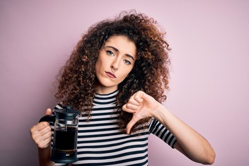 Young beautiful woman with curly hair and piercing doing coffee holding french coffeemaker with angry face, negative sign showing dislike with thumbs down, rejection concept