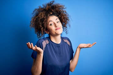 Young beautiful woman with curly hair and piercing wearing casual blue t-shirt clueless and...